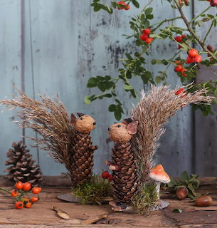 Two handmade squirrel figures crafted from pine cones and dried grasses with rose hips and mushroom on wooden surface