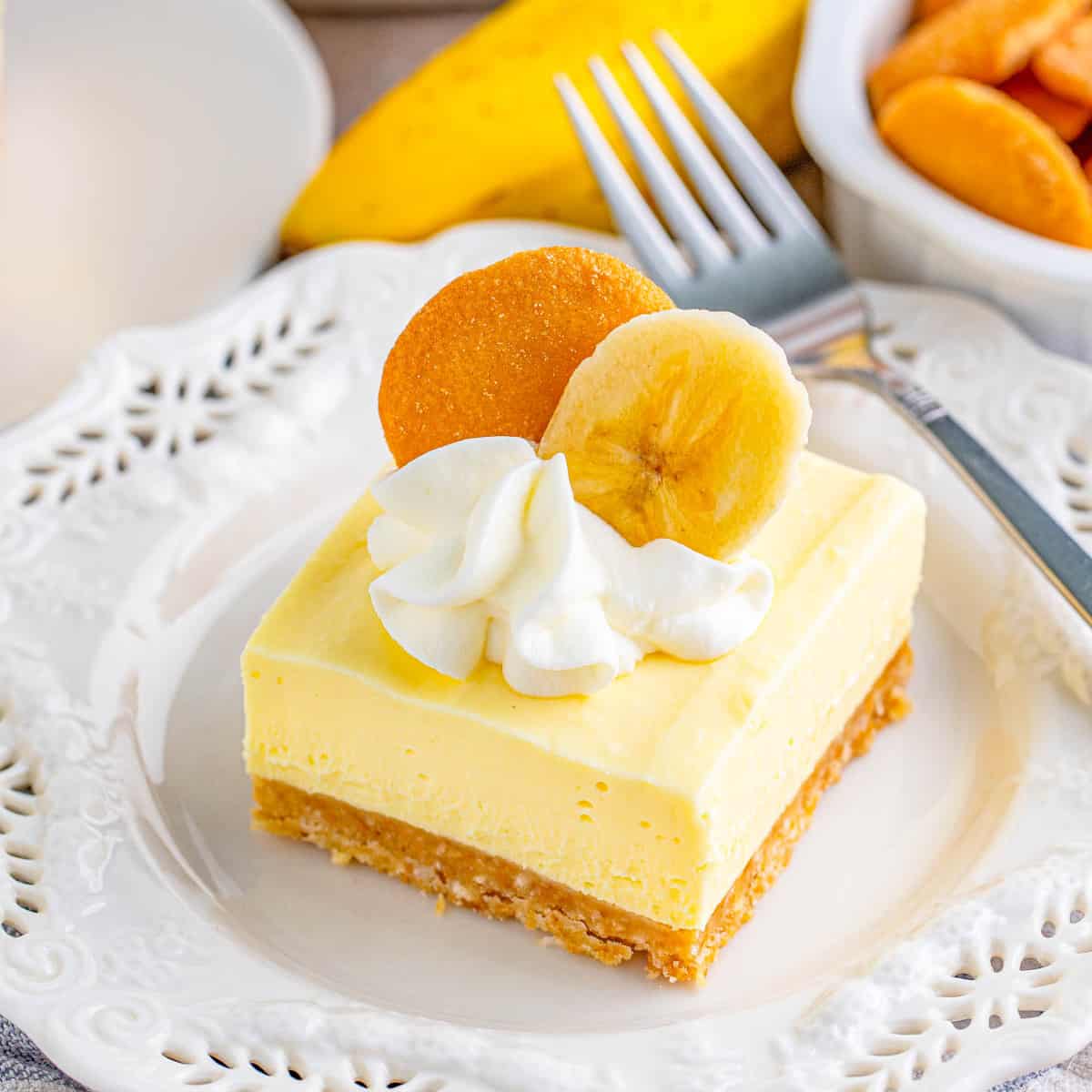 Square banana cheesecake bar with whipped cream, banana slice, and cookie on white plate