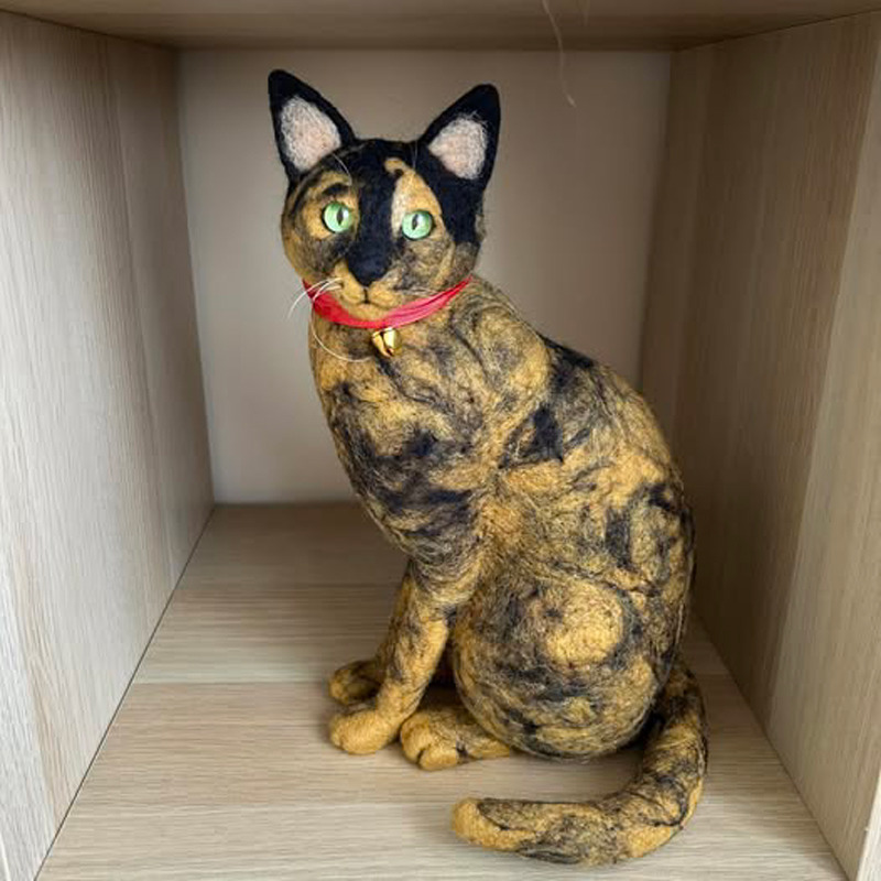 Tortoiseshell cat sculpture with green eyes and red collar sitting on a wooden shelf