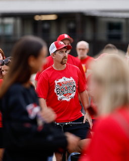 Man in red shirt and white cap looking nervous among crowd near a bus at a softball event