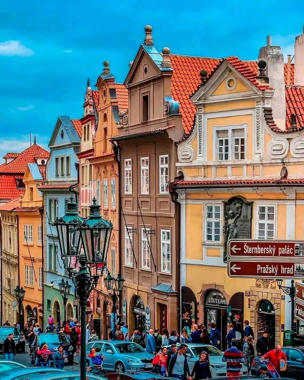 Colorful historic buildings with ornate facades and red roofs on a busy Prague street with pedestrians and blue sky