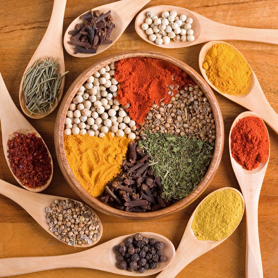 Wooden bowl containing colorful spices and seeds arranged in sections, surrounded by wooden spoons with various spices