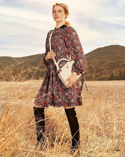 Woman in a patterned dress and black boots standing in a dry field with hills behind her