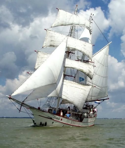 White tall ship with multiple white sails on water against cloudy blue sky