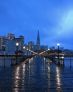 Wet Pier 7 boardwalk at dusk with lit lampposts and San Francisco Financial District skyline