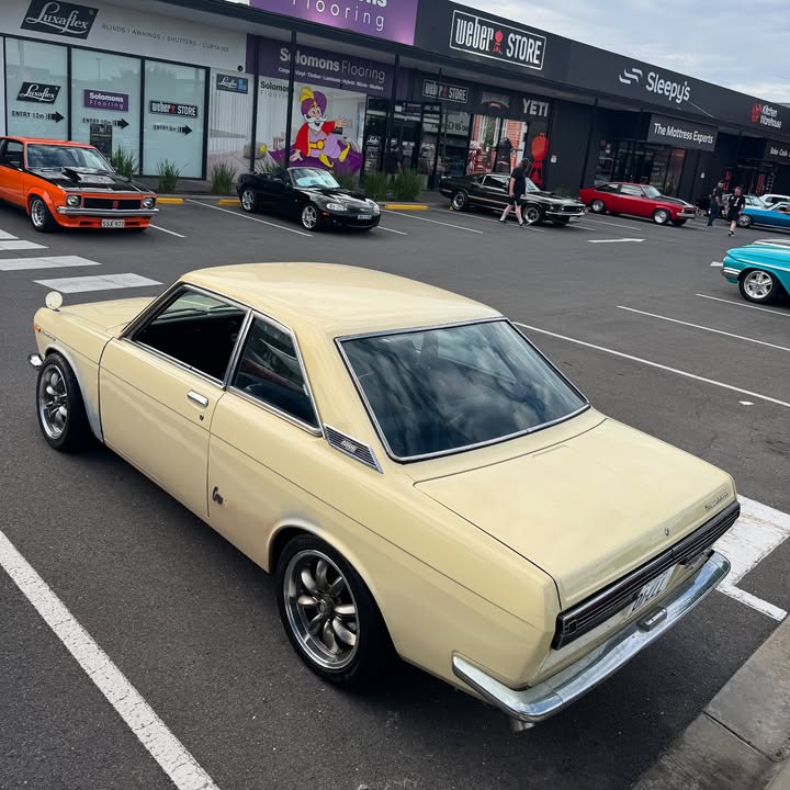 Cream-colored classic coupe parked on a street with vintage cars and shops behind it