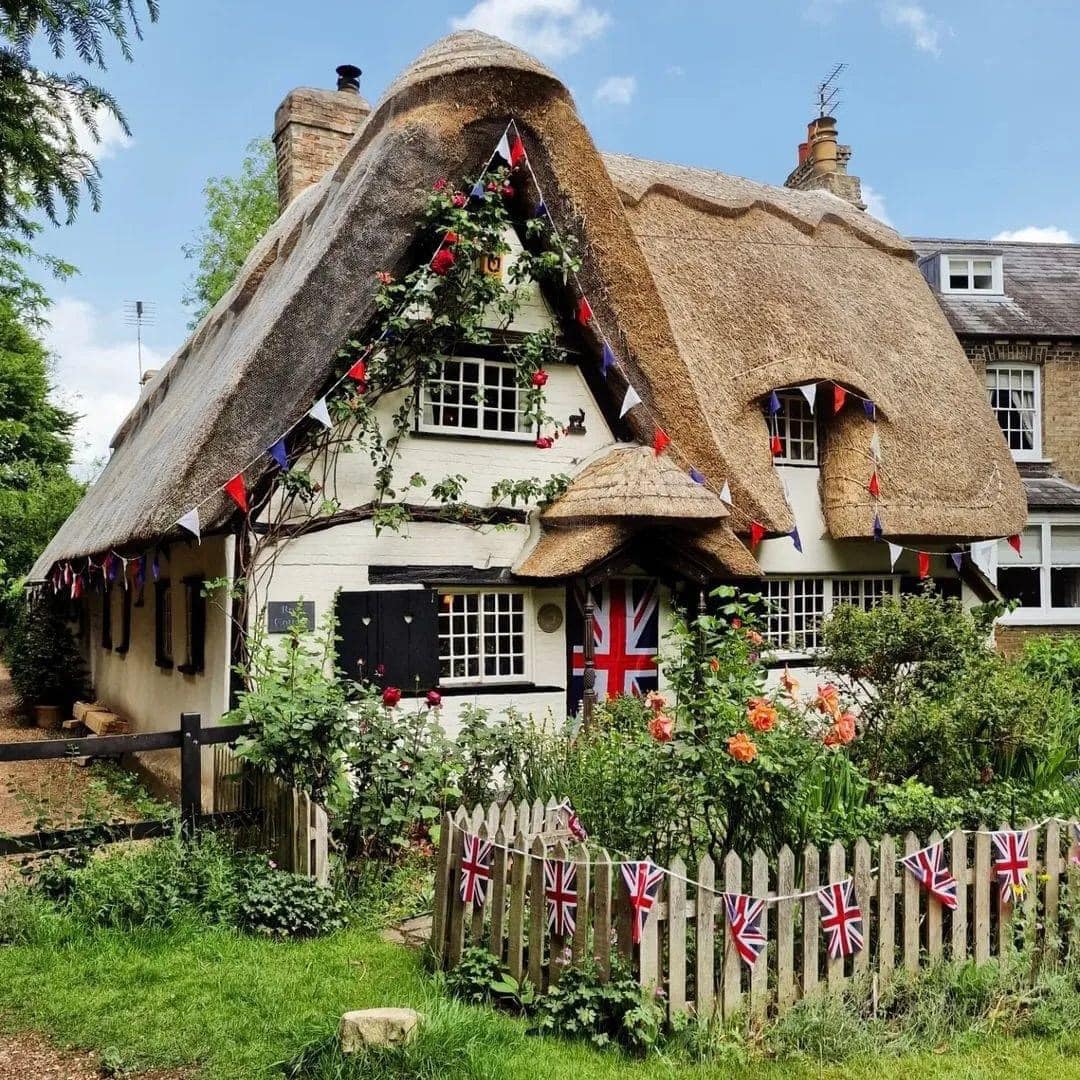Traditional English cottage with thatched roof, white walls, roses climbing facade, surrounded by garden and picket fence