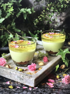 Two layered pistachio desserts in glass cups topped with crushed pistachios and pink petals on marble tray