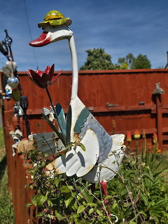 White swan garden ornament with a small hat among plants by a red wooden fence
