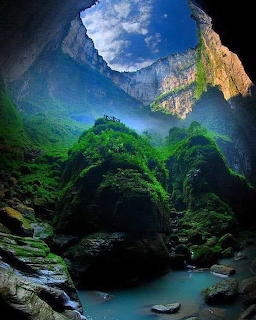 View from inside a giant sinkhole cave with green mountains, blue pool, and sky opening above