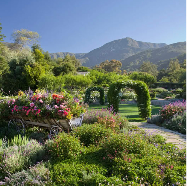 Lush garden with flowers, a vine-covered archway, and mountains in the background