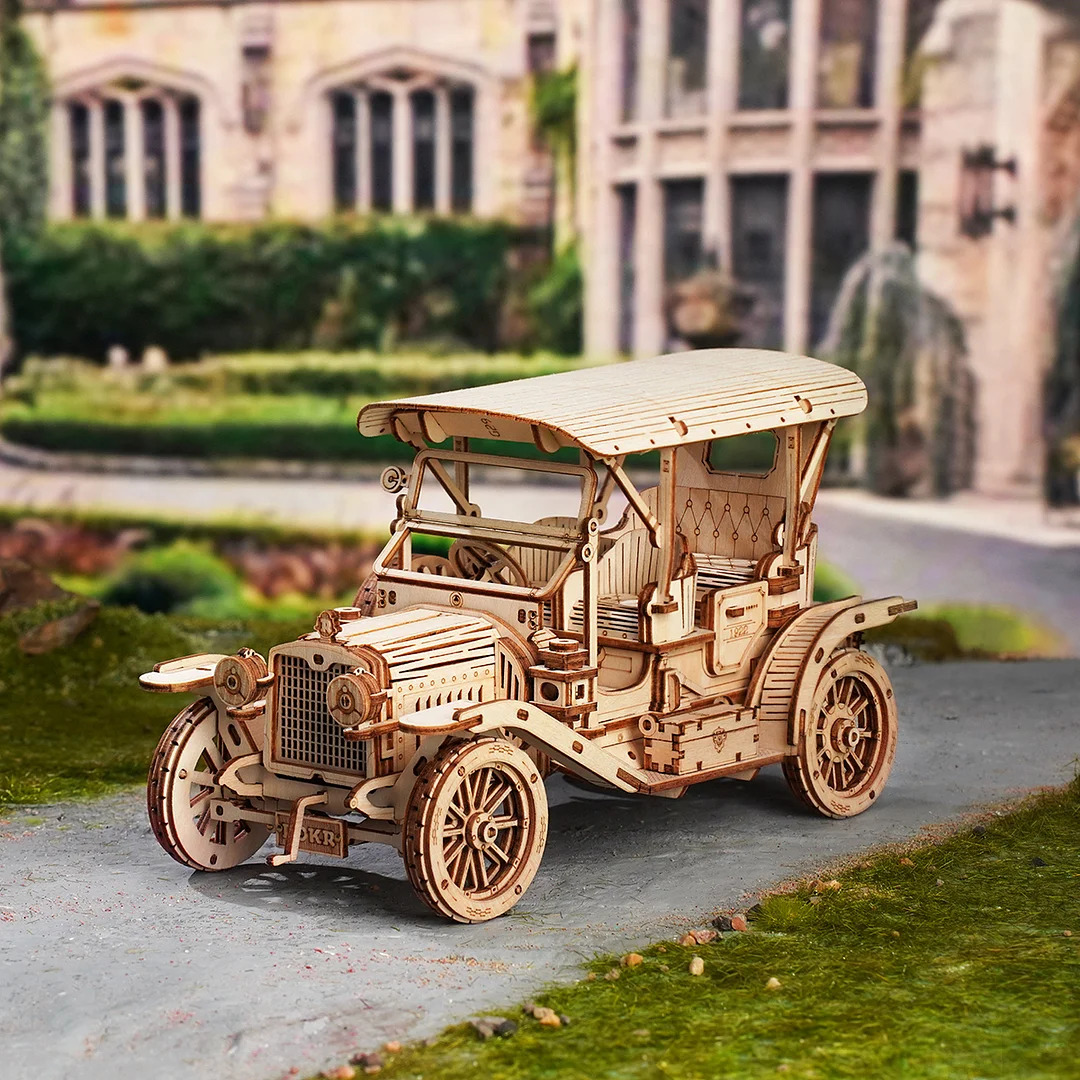 Wooden puzzle model of vintage car with canopy on grass, historic building behind