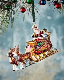 Glass Santa sleigh ornament hanging from pine branches with colorful baubles and soft bokeh background