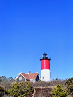 Red and white lighthouse next to a small house on dunes beneath a bright blue sky.
