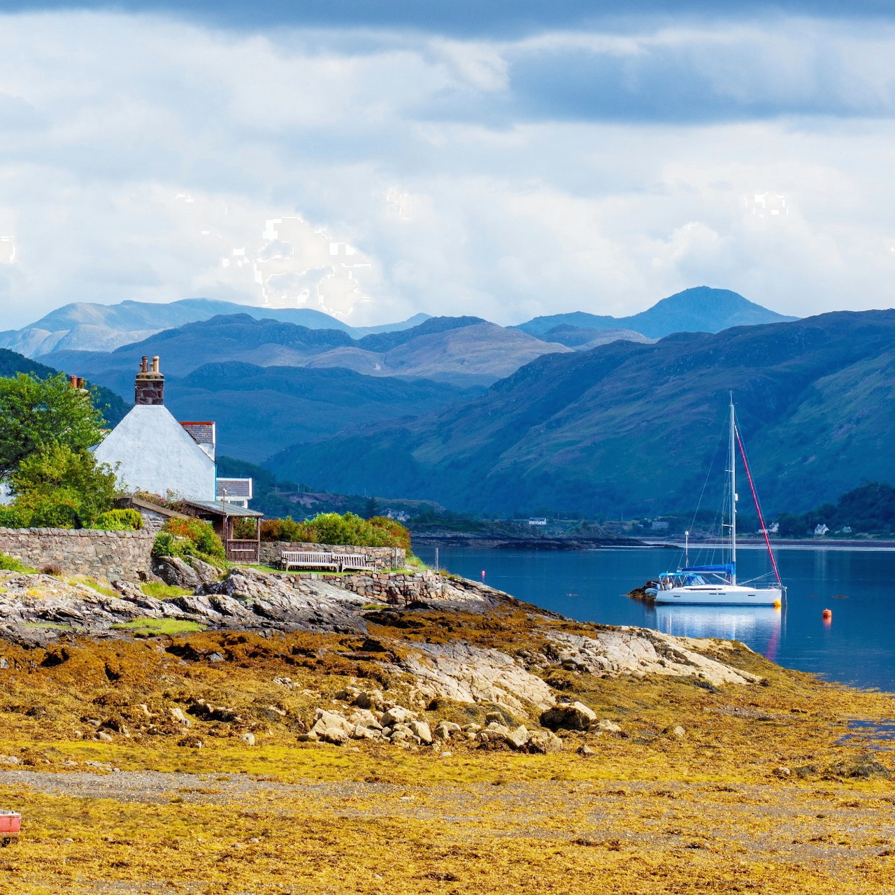 White cottage on a rocky Scottish coast with a sailboat and blue mountains in the background