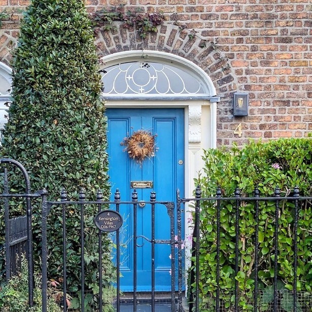 Blue front door with wreath in a brick wall, bordered by shrubs and a black iron fence.