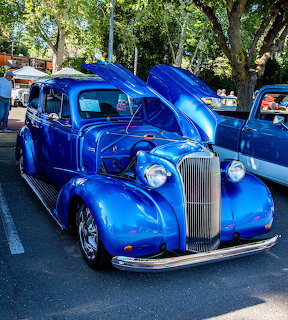 Shiny blue vintage car with open hood at an outdoor car show surrounded by trees