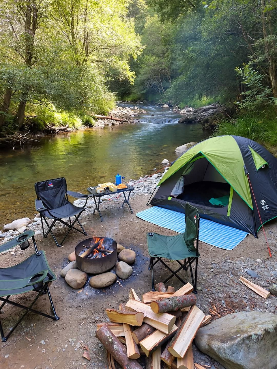 Green tent and campfire with chairs beside a forest river campsite