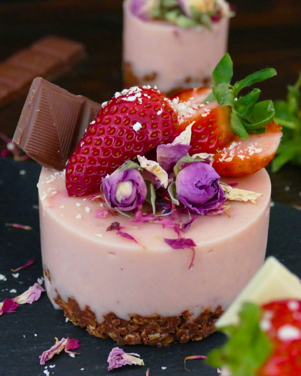 Pink strawberry cheesecake topped with a strawberry, purple roses, and chocolate on slate