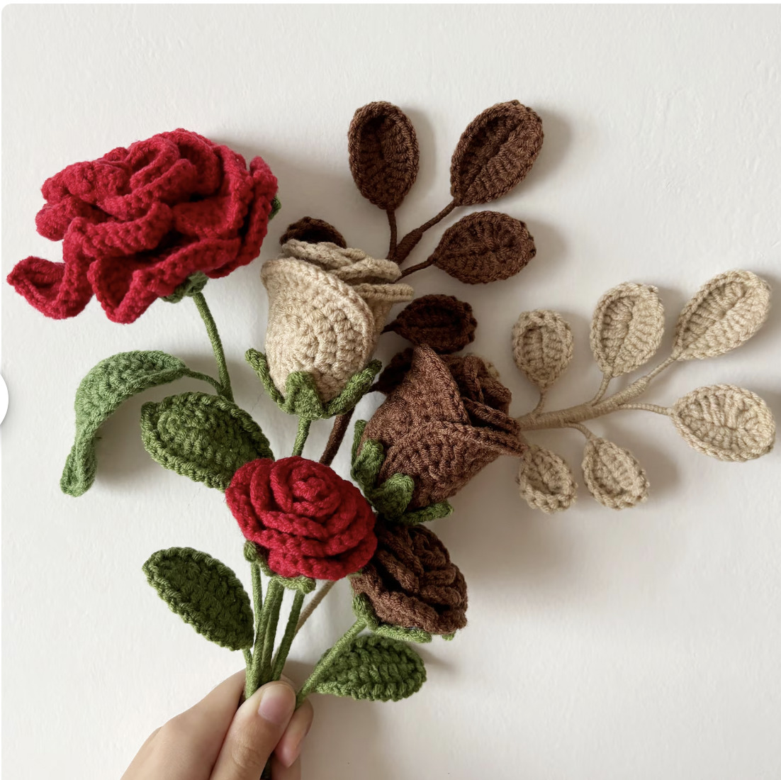 Hand holding crocheted roses in red and cream with green leaves and brown yarn pieces on white background