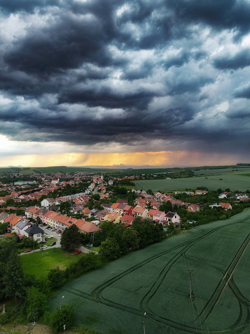 Aerial view of Hluk village with red rooftops and green fields under dark storm clouds at dusk.