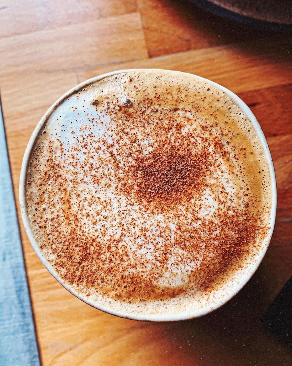 Top-down view of a frothy latte with brown powder dusting in a white cup on wood surface