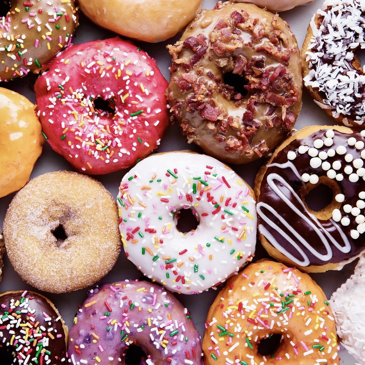 Overhead view of assorted colorful donuts with icing, sprinkles, coconut, chocolate drizzle, and bacon.