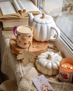 Autumn tea arrangement with white pumpkins, floral mug, and open book on windowsill with fall leaves