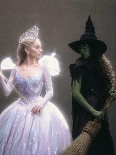 Glinda in a sparkling gown stands beside green-skinned Elphaba in black with a witch hat