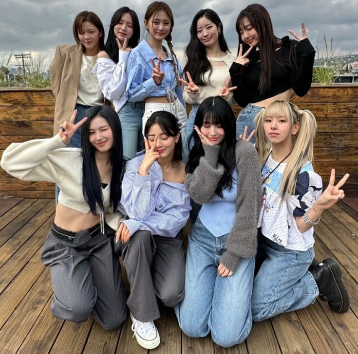 Eight young women posing together on a wooden deck, making peace signs under a cloudy sky