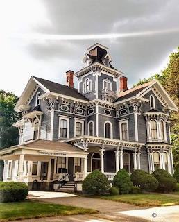 Gray Victorian mansion with white trim, cupola tower, and front porch on lawn under stormy sky