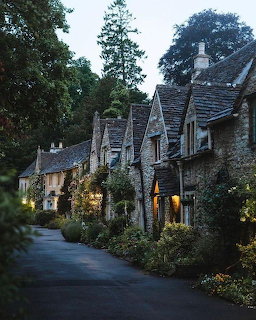 Row of historic stone cottages along a quiet lane at dusk with glowing windows and tall trees