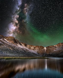 Milky Way and green aurora over mountains reflected in a calm lake at night