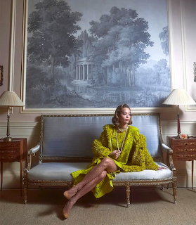 Woman in bright yellow feathered coat sitting on gray sofa against painted landscape mural backdrop