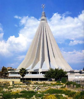 Tall conical sanctuary church with ribed white walls and a spire topped by a statue, against blue sky
