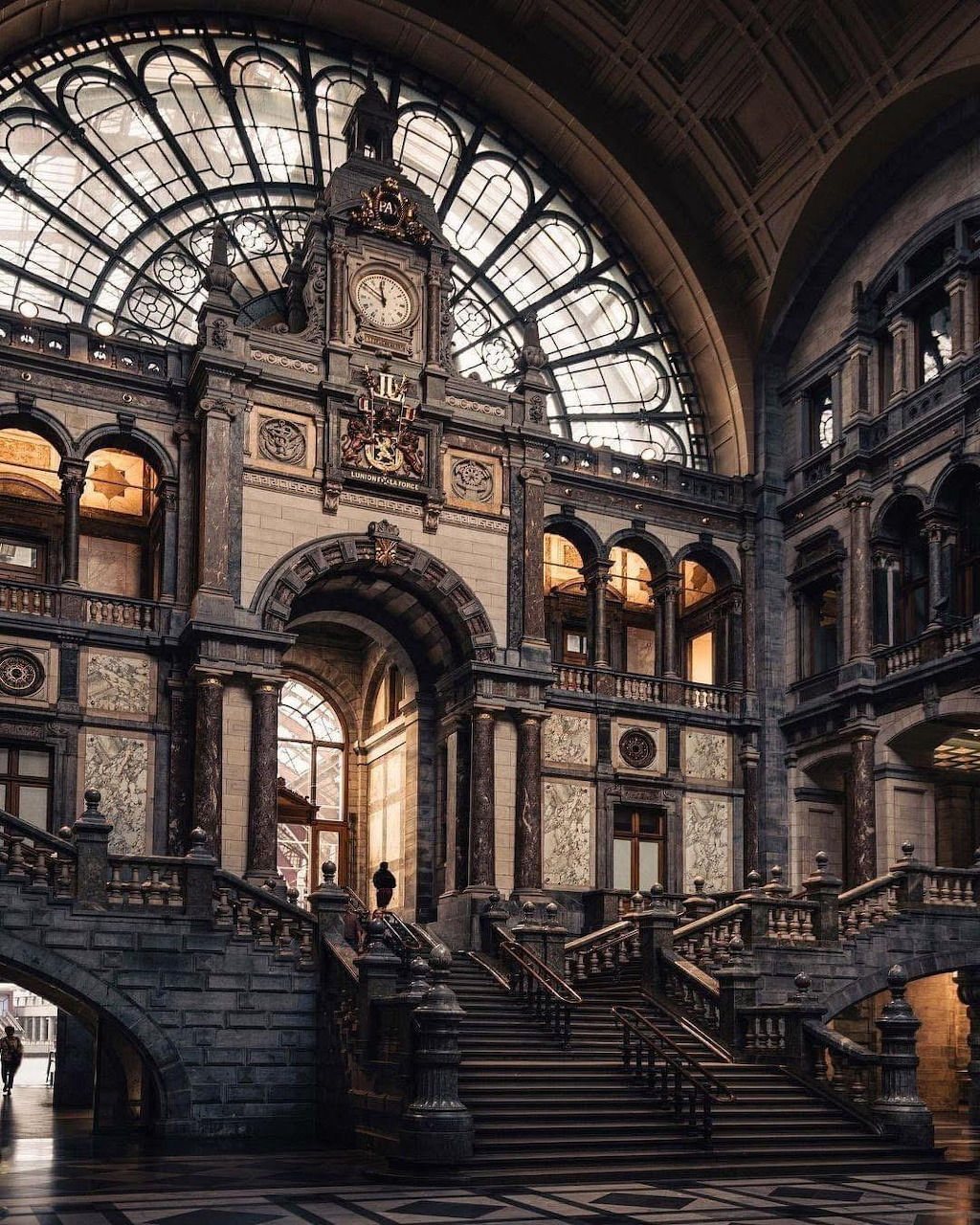 Ornate interior hall of Antwerp Central Station with vaulted glass ceiling, grand stone archway and twin staircases