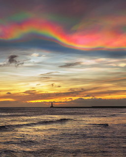 Rainbow-like cloud over the ocean at sunset with waves and a distant lighthouse