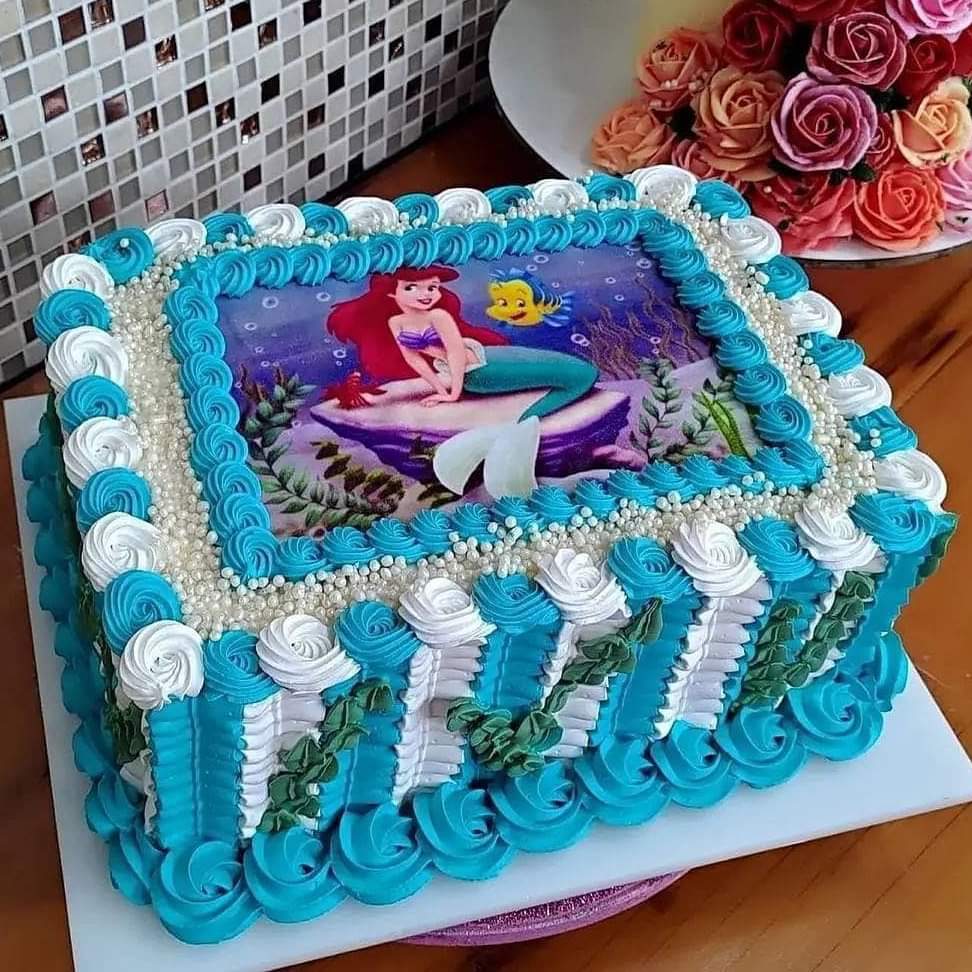 Blue frosted cake with an Ariel and Flounder picture, white icing swirls, and roses nearby