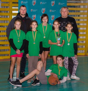 Girls in green shirts with medals and a trophy posing with two adults in a gym