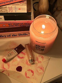Lit pink jar candle next to stacked books, lipstick, perfume bottle, and napkin with red kiss marks