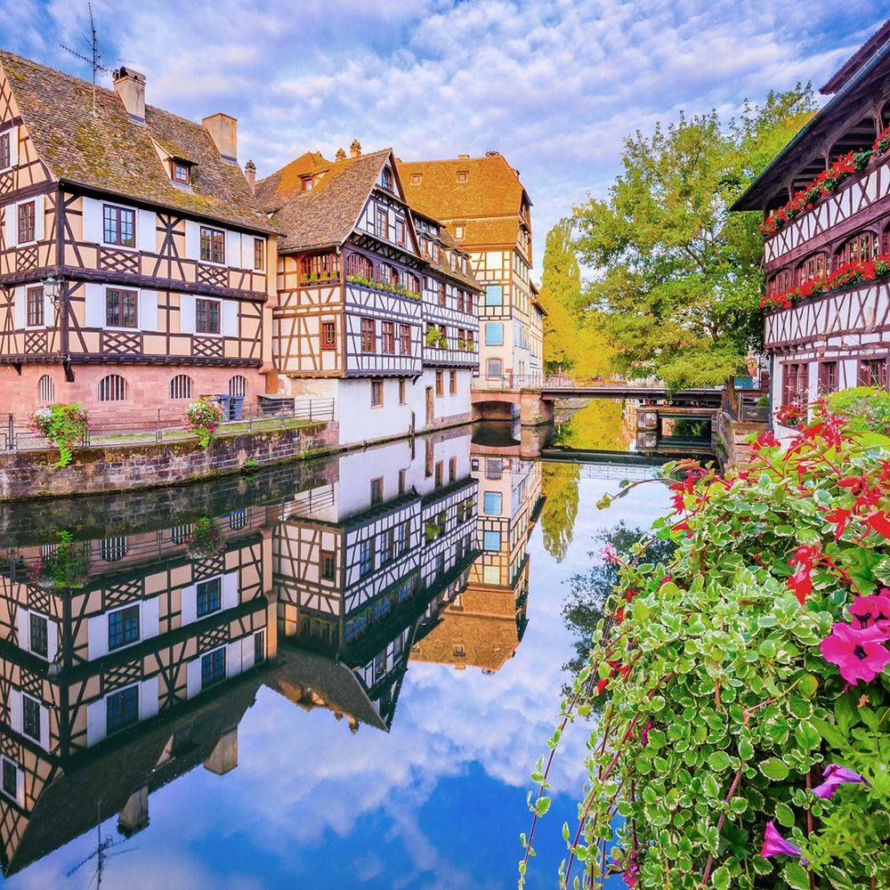 Half-timbered houses reflected in a calm canal with flowers and a small bridge