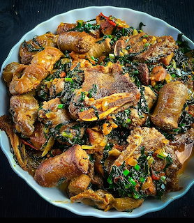 White bowl of African pork stew with meat pieces, leafy greens, and vegetables on a dark table