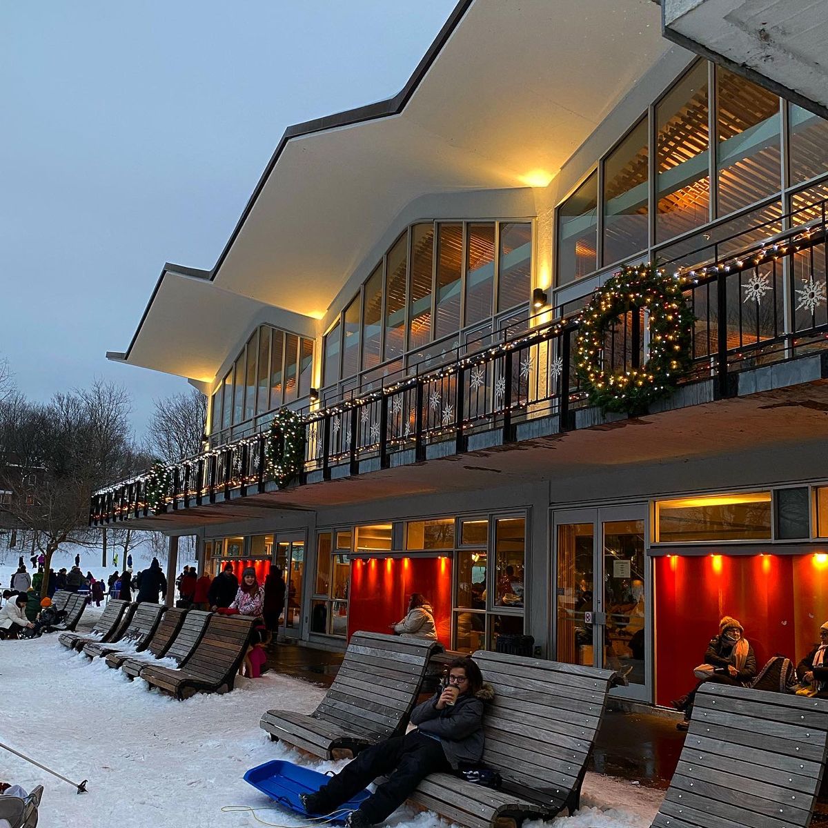 Illuminated modern pavilion with curved roof and glass walls at twilight, people on lounge chairs in snow
