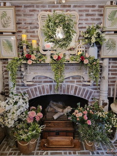 Brick fireplace mantel decorated with floral garlands, roses, greenery wreath, candles, and botanical framed prints