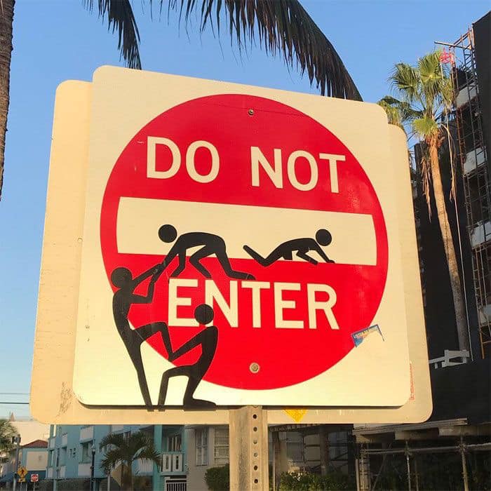 Modified do not enter street sign with stick-figure graffiti, palm trees, and buildings