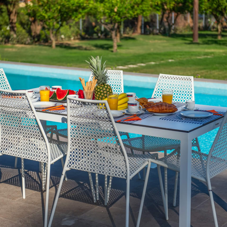 Outdoor patio table and chairs beside a swimming pool, set with fruit and drinks