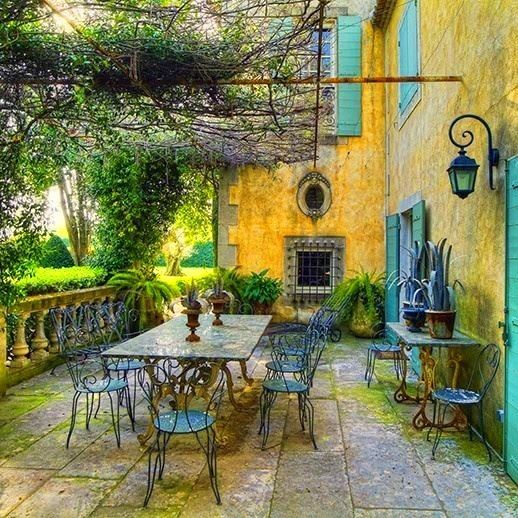 Sunlit European patio with iron table and chairs beside a yellow house with blue shutters and climbing vines