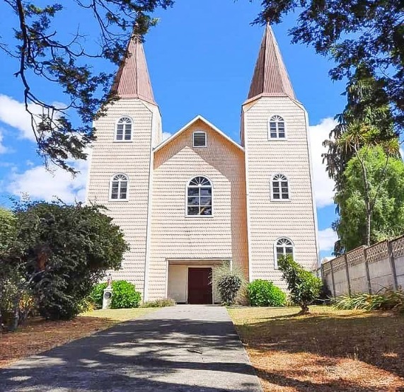 White wooden church with twin steeples and a central entrance, surrounded by trees and blue sky