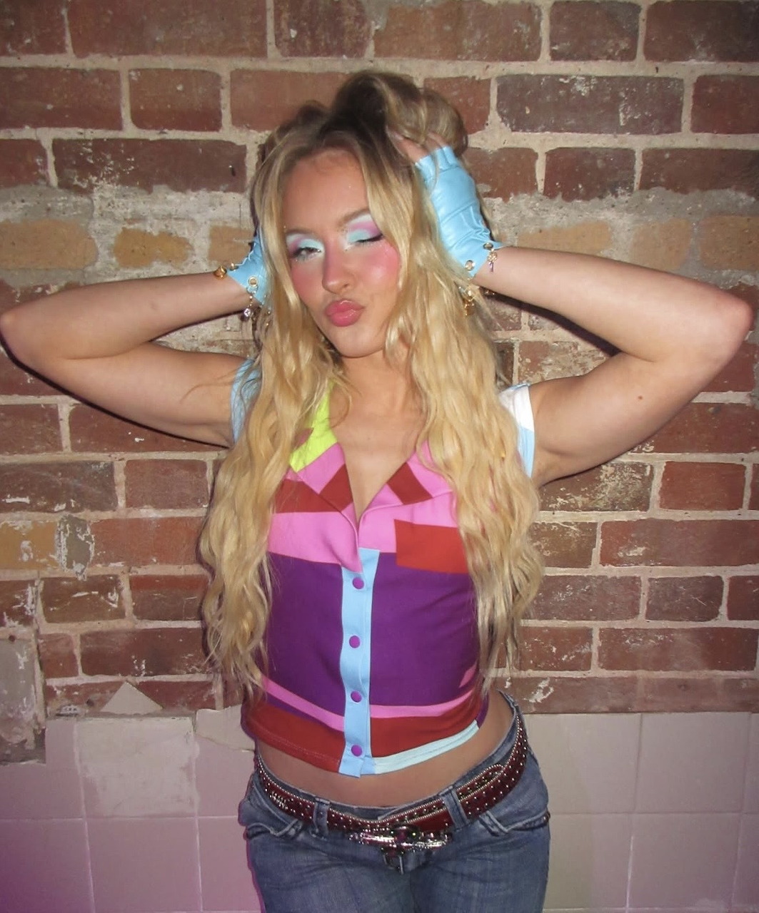 Woman in colorful Y2K outfit with blue hair accessories posing against brick wall
