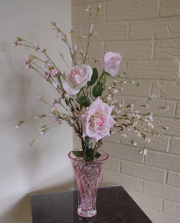 Pink roses and small blossoms arranged in a pink glass vase on a dark table near a white brick wall
